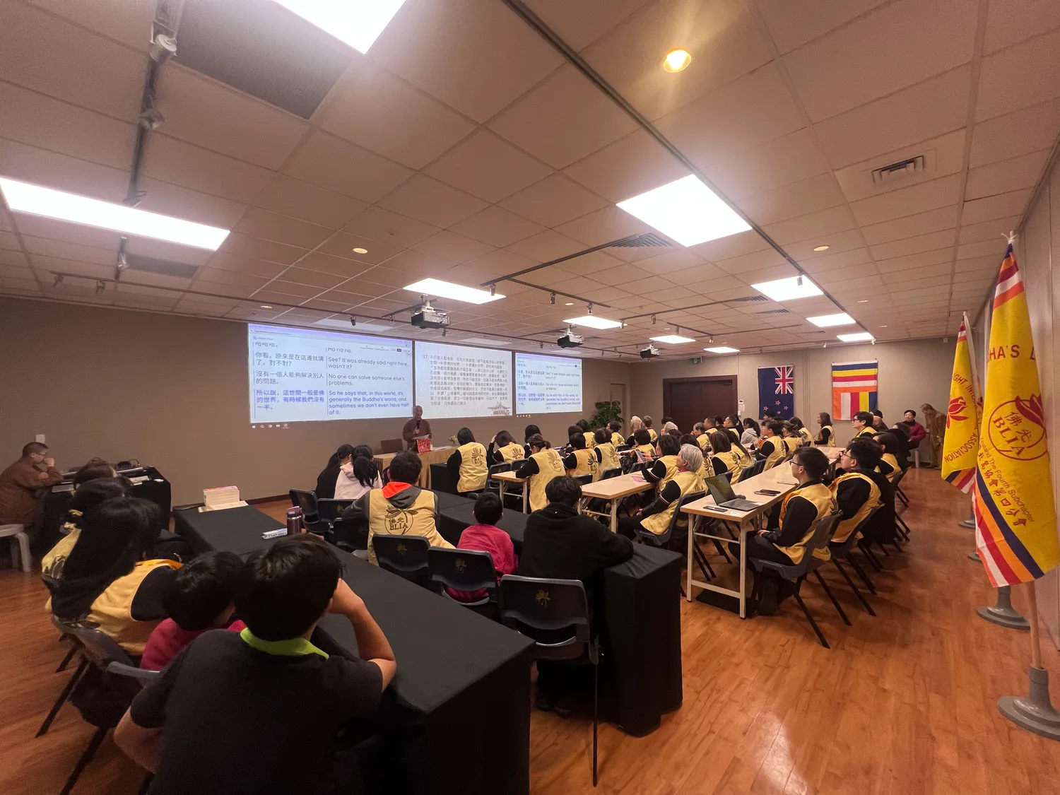 Members in BLIA vests follow a teaching at an overseas chapter, with Orato Flow’s Mandarin and English captions on the screens.