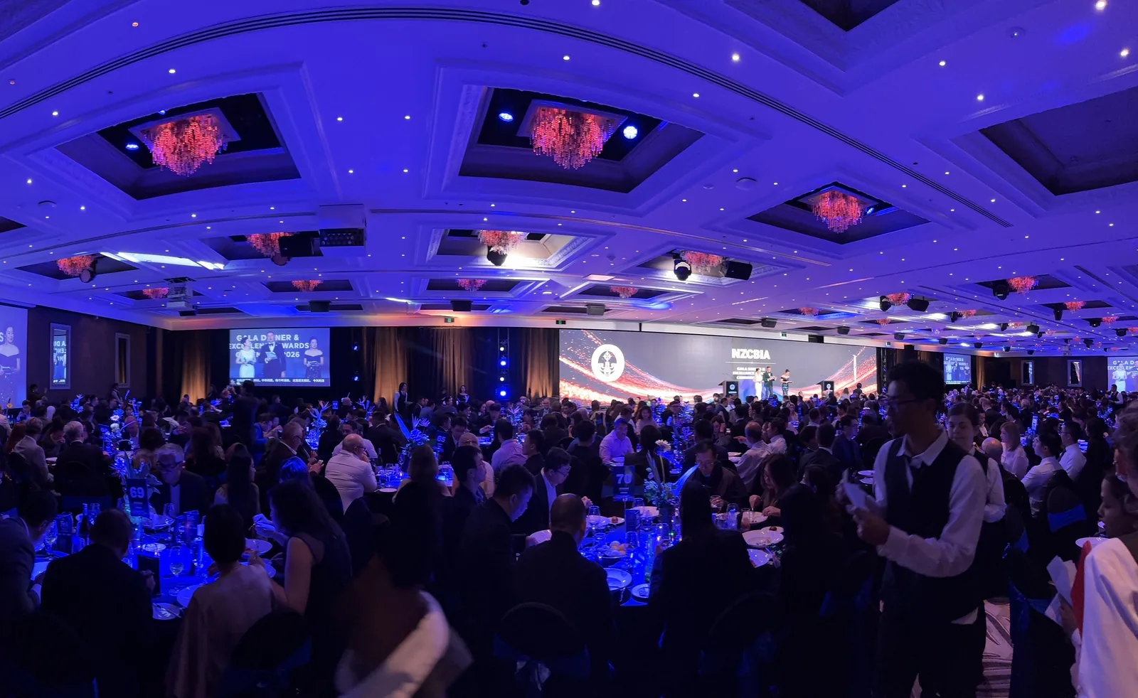 The Great Room at Cordis Auckland filled with 850 guests under blue lighting at the 2026 NZIBES Gala Dinner, with the stage and screens at the far end.