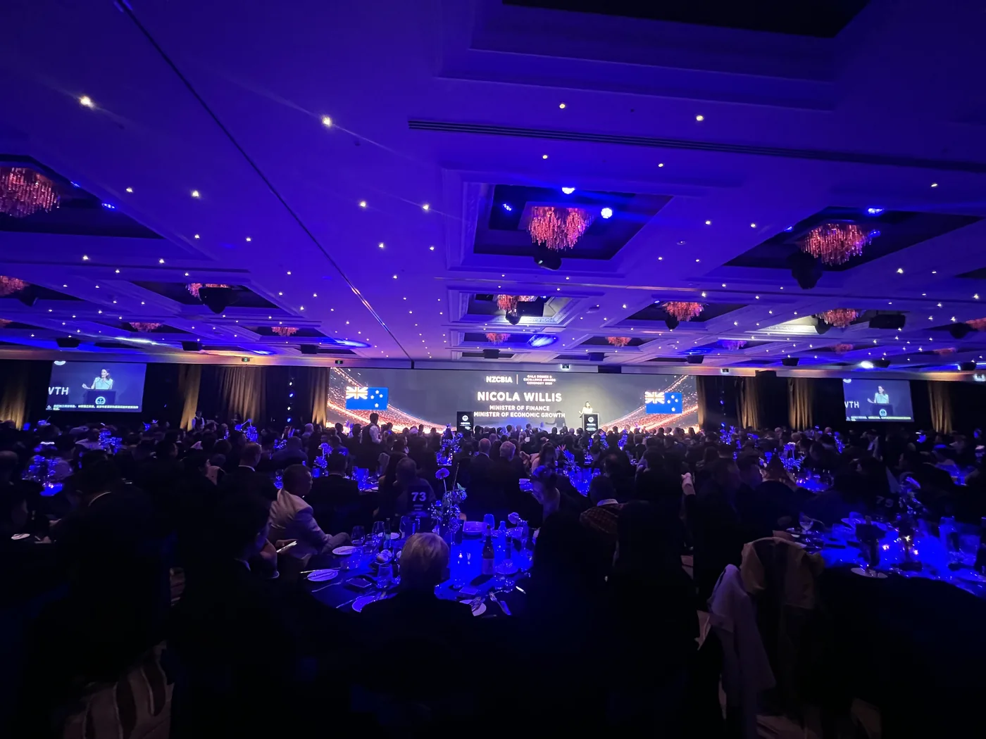 A wide view of the 2026 NZIBES Gala Dinner as Nicola Willis, Minister of Finance, is introduced on the main stage screen, with live captions on the side screens.