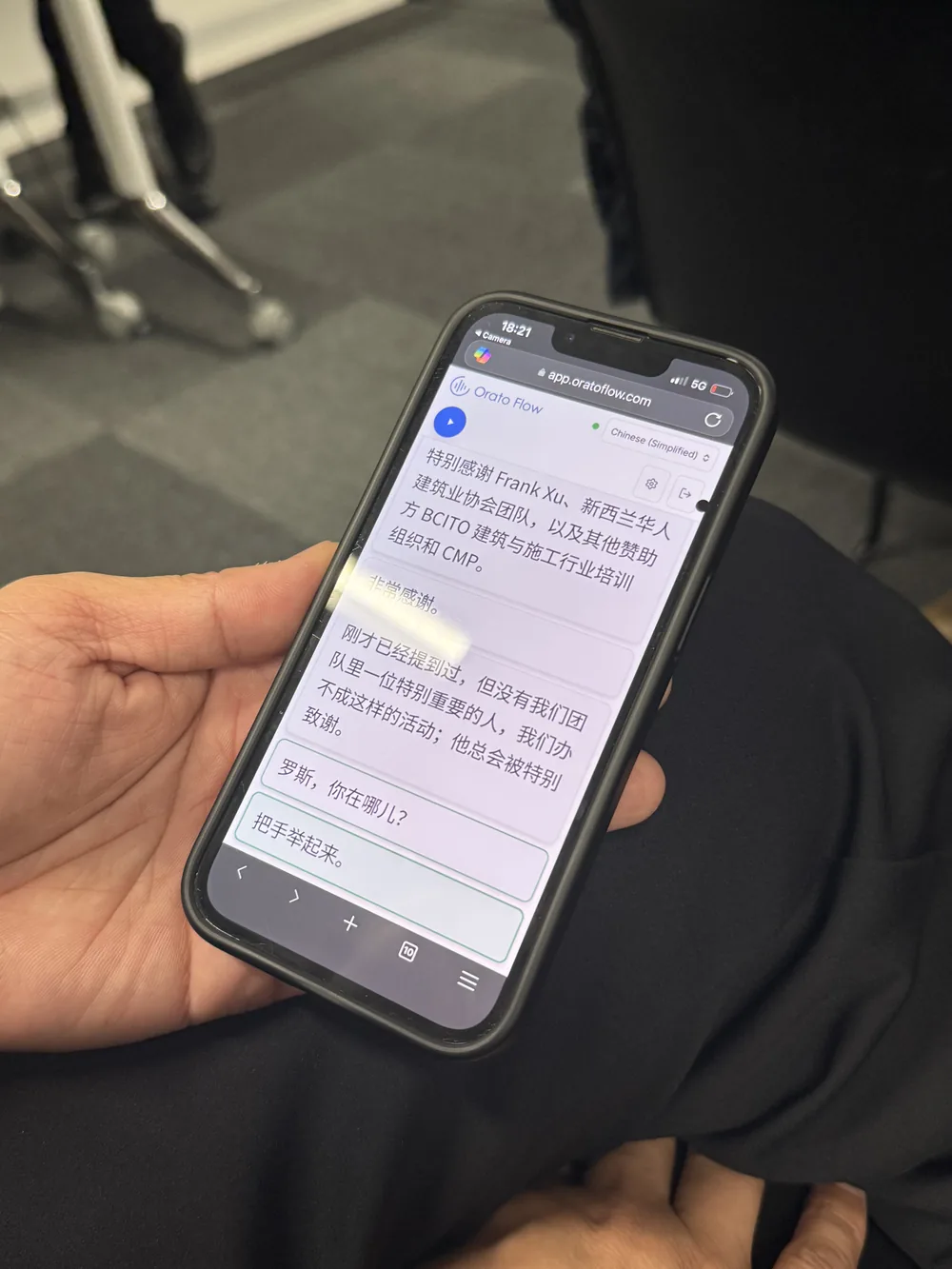 A hand holding a phone showing Orato Flow open in a mobile browser, with the live Chinese translation of the talk and a Chinese (Simplified) language selector at the top.
