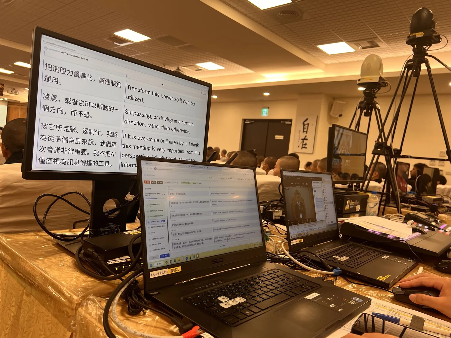 A monitor showing Orato Flow’s side-by-side Chinese and English transcript at the symposium, beside the AV operator’s laptops.